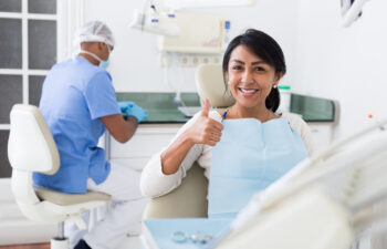 A smiling female patient sits in a dental chair, giving a thumbs-up to the camera. In the background, a male dental professional in blue scrubs and a face mask is preparing the treatment area in a bright, modern dental office.