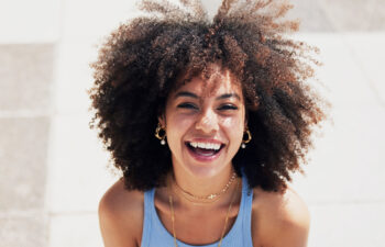 A young woman with curly hair smiling brightly outdoors, wearing gold jewelry and a light blue tank top.