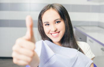 A satisfied young woman sitting in a dental chair, wearing a dental bib, smiles and gives a thumbs up to the camera.